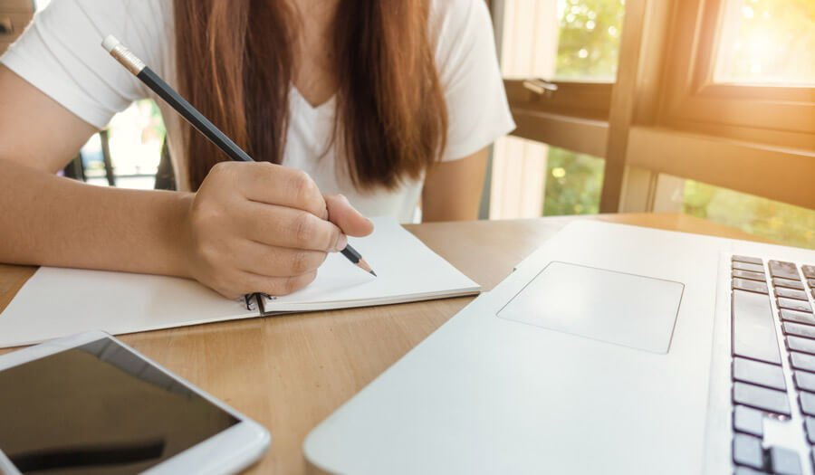 Uma mulher segurando um lápis em cima de um caderno, observando em segundo plano um notebook